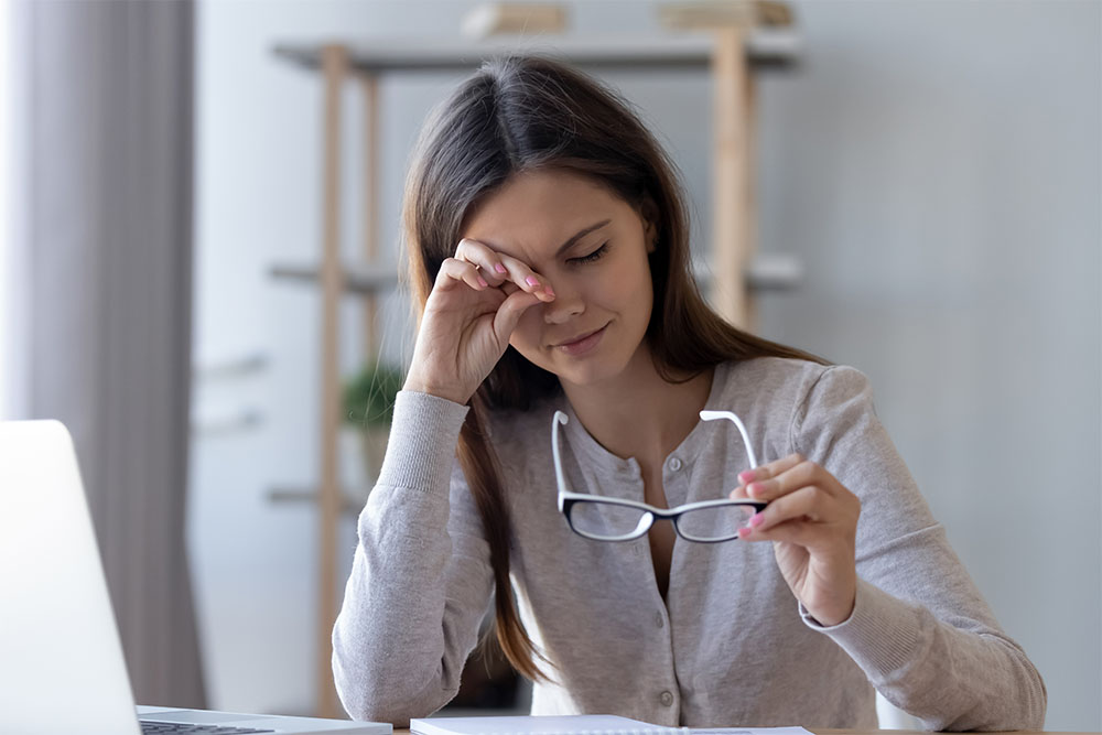 Donna al computer che si stropiccia gli occhi stanchi tenendo in mano gli occhiali, sintomo di astigmatismo elevato.